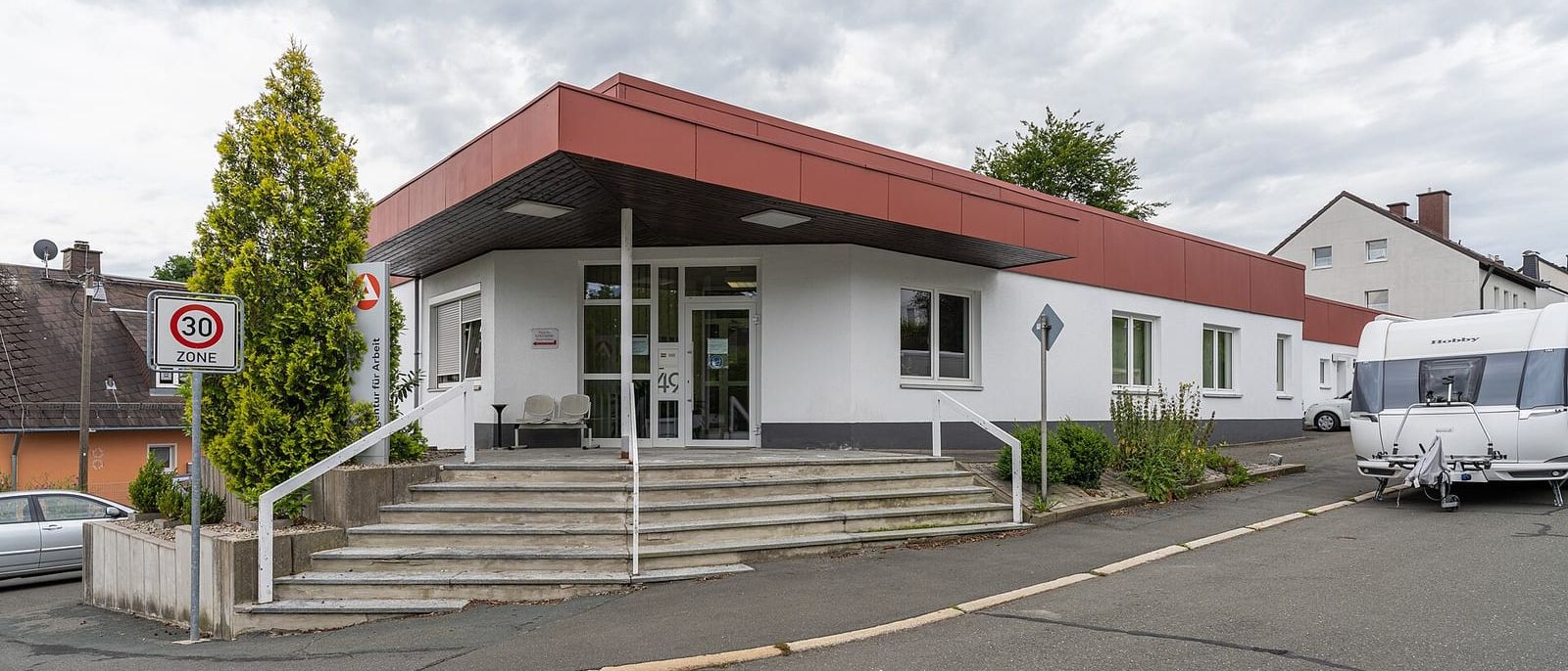Flaches Gebäude der Agentur für Arbeit mit rotem Dachrand, Treppe und Eingangsschild