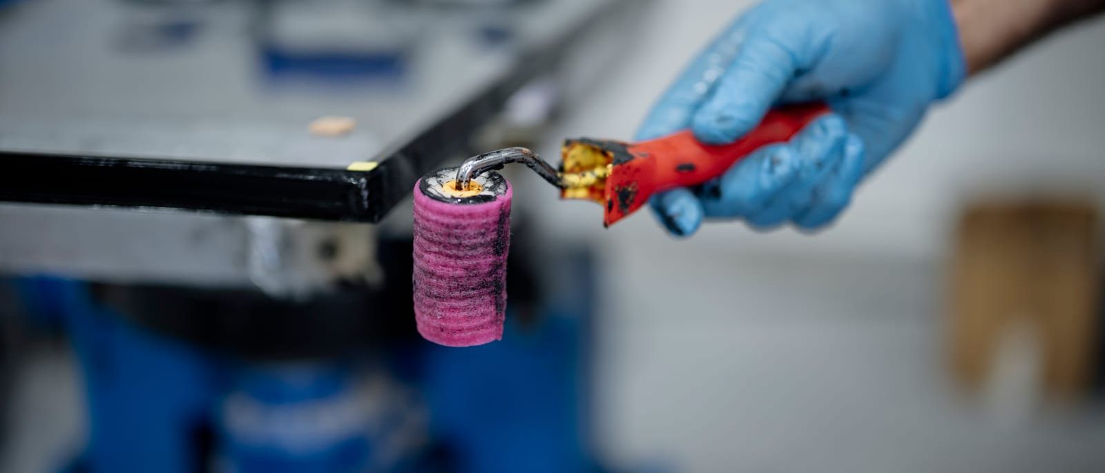 Hand im blauen Handschuh hält eine gebrauchte Farbrolle in einer Werkstatt
