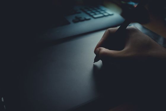 Hand mit Eingabestift auf einem Grafiktablett vor einer Tastatur in gedämpftem Licht