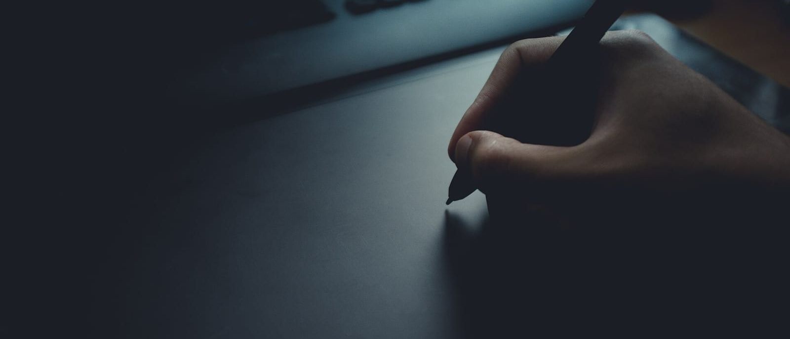 Hand mit Eingabestift auf einem Grafiktablett vor einer Tastatur in gedämpftem Licht