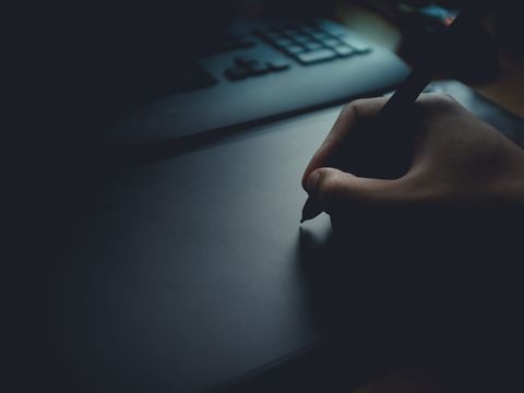 Hand mit Eingabestift auf einem Grafiktablett vor einer Tastatur in gedämpftem Licht