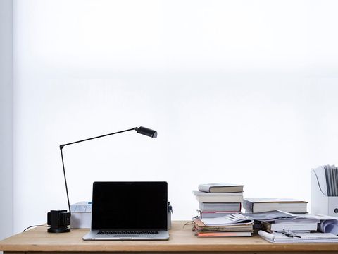 Aufgeklappter Laptop, schwarze Schreibtischlampe und Bücherstapel auf einem Holztisch vor weißer Wand