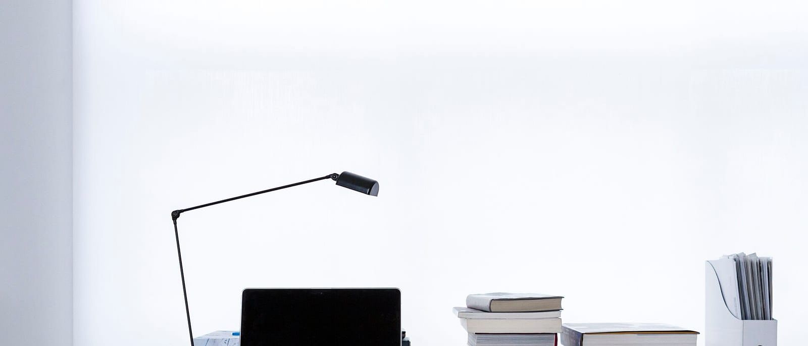 Aufgeklappter Laptop, schwarze Schreibtischlampe und Bücherstapel auf einem Holztisch vor weißer Wand