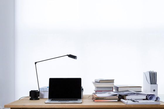 Aufgeklappter Laptop, schwarze Schreibtischlampe und Bücherstapel auf einem Holztisch vor weißer Wand